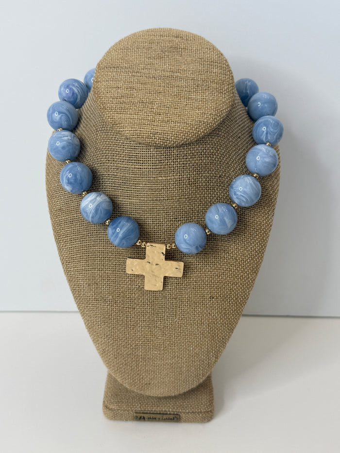 The Skyline Necklace in periwinkle blue with hammered gold cross pendant on a linen jewelry bust