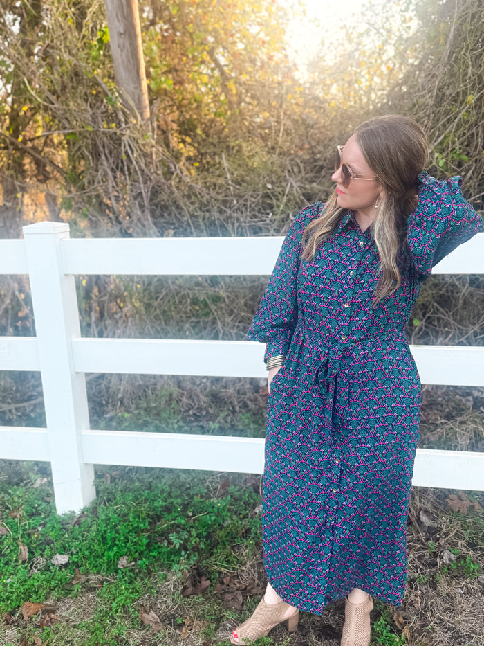 Woman in a blue floral dress standing in front of a white picket fence with trees in the background.