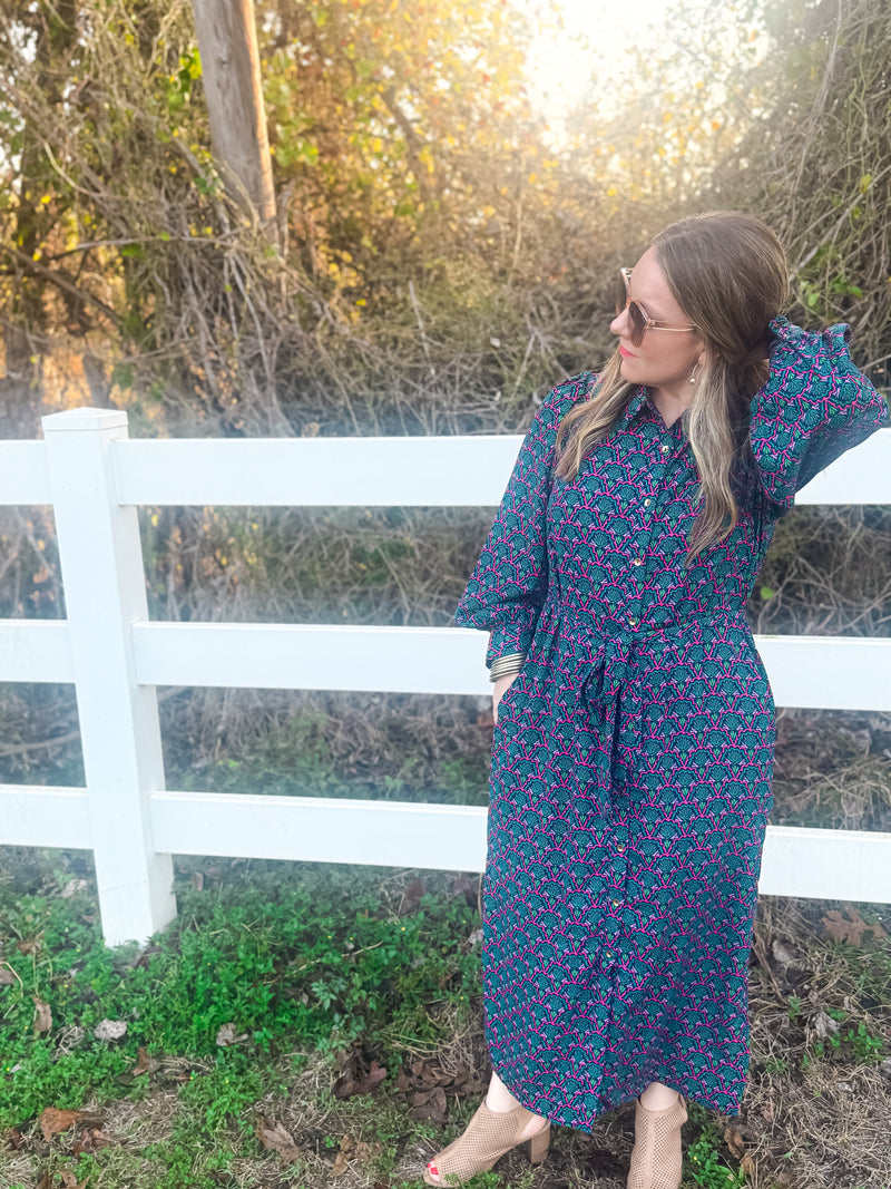 Woman in a blue floral dress standing in front of a white picket fence with trees in the background.