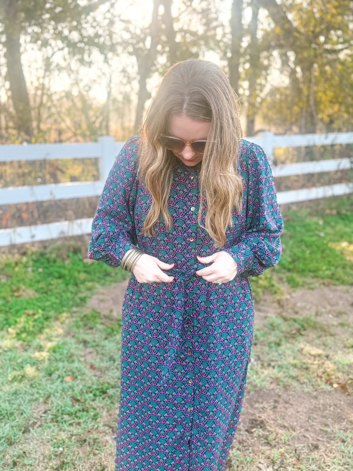 Woman in a floral dress standing in a field with a white fence and trees in the background